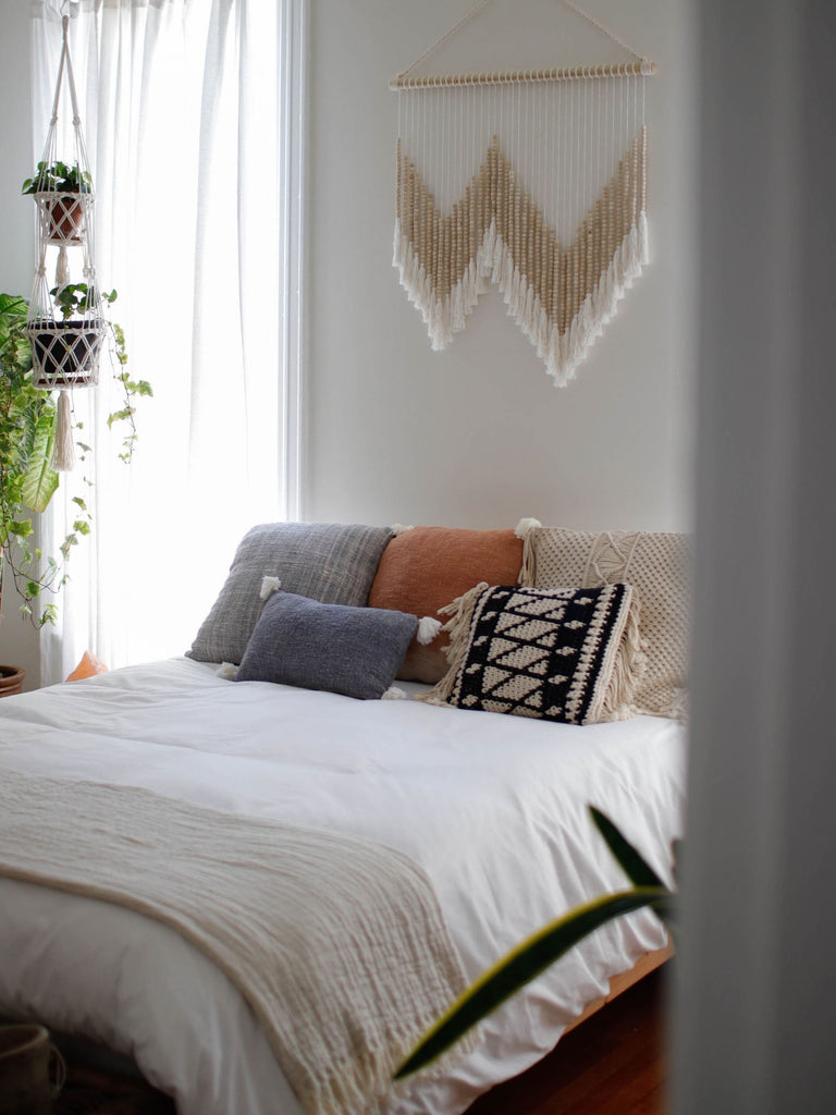 handmade beaded wall hanging art and macrame plant hangers above a bed in a light filled bohemian bedroom