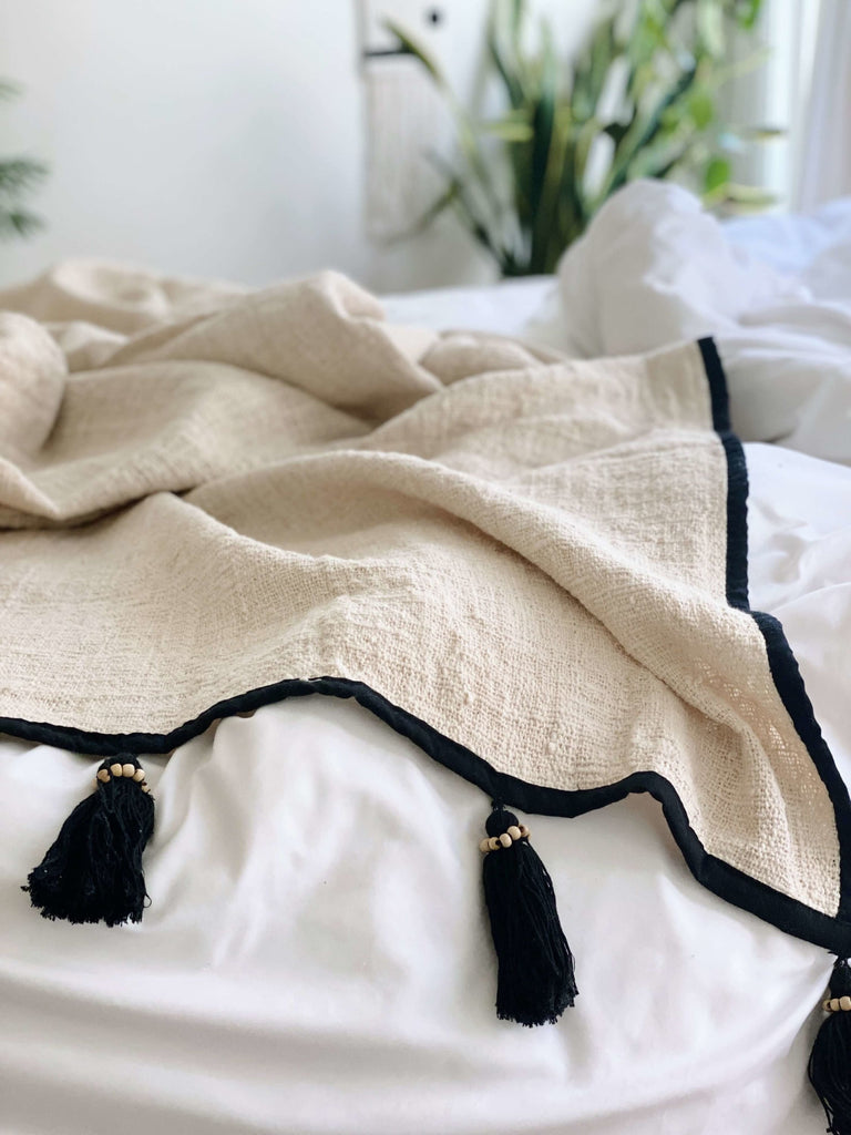 beautiful bohemian handwoven jute and linen cream cozy throw blanket with black tassels on a white bean bag chair