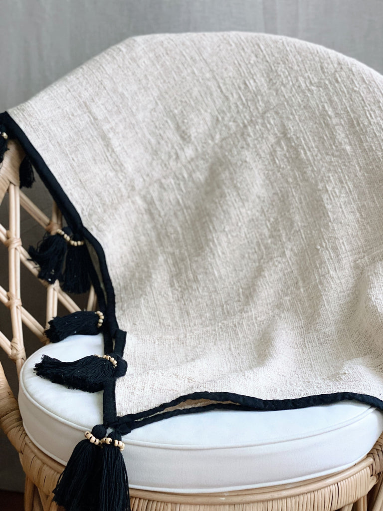 scandi bohemian jute and linen throw blanket with black tassels over a wicker chair