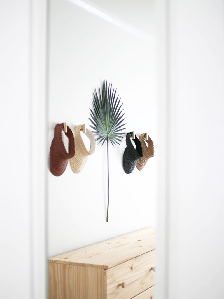 reflection of ethereal bohemian decor using handwoven palm leaf sun visors hanging on gold hooks with a large green palm leaf