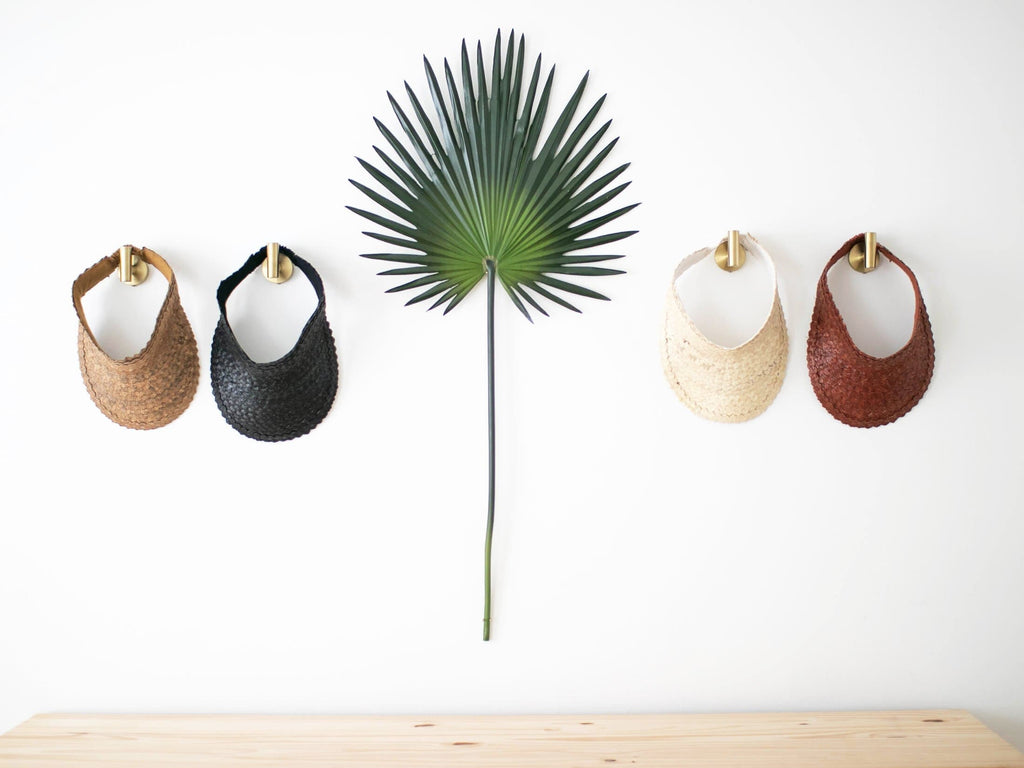 bohemian decor using handwoven palm leaf sun visors hanging on gold hooks with a large green palm leaf in light filled room