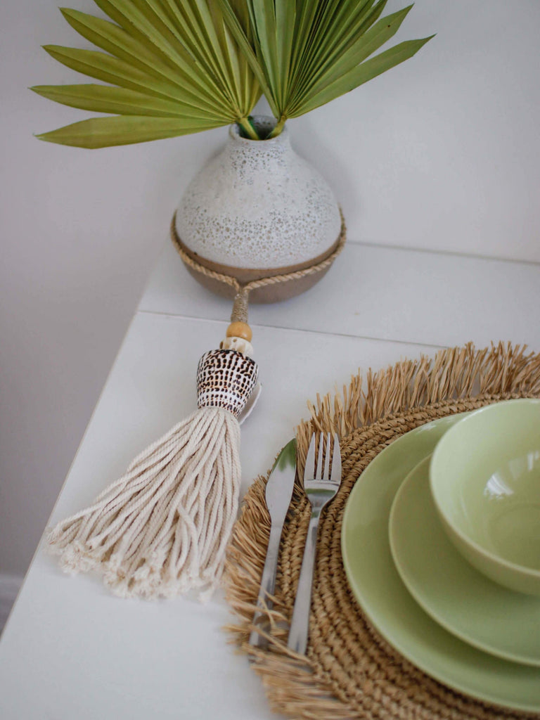 dining table setup with handwoven natural fringe placemat and sage plates next to a pot with tassel and palm leaves