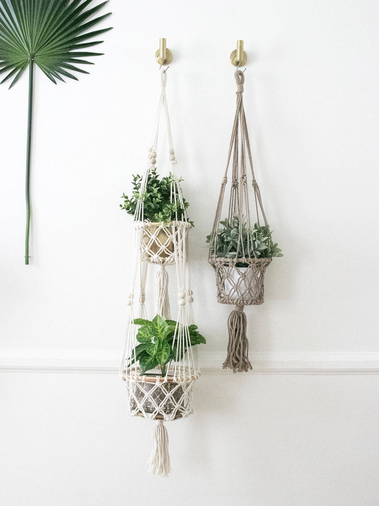 cream macrame double plant hanger and single brown macrame plant hanger holding beautiful plants on a white wall
