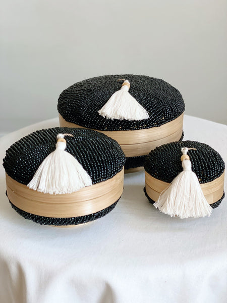set of three round hand-beaded black bamboo baskets with cream tassel on white table