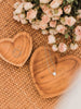 two hand-carved heart teak dishes with dainty silver jewelry and dried pink roses on a wicker table