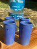 closeup of four blue handmade high fire clay shot glasses on a wooden board with a bottle of cupreata