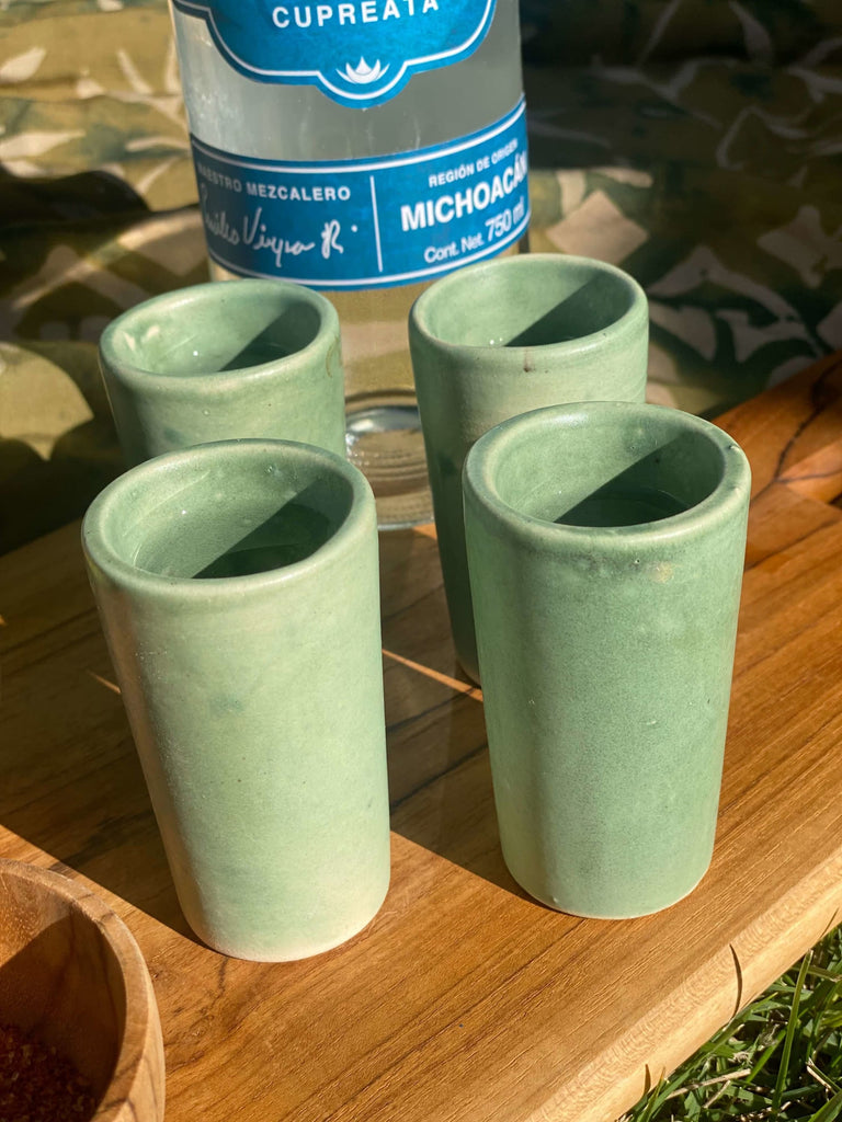closeup of four green handmade high fire clay shot glasses on a wooden board with a bottle of cupreata