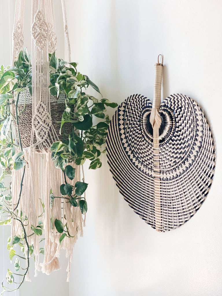 large natural black bohemian handwoven palm leaf fan hanging on white wall next to macrame plant hanger with pothos