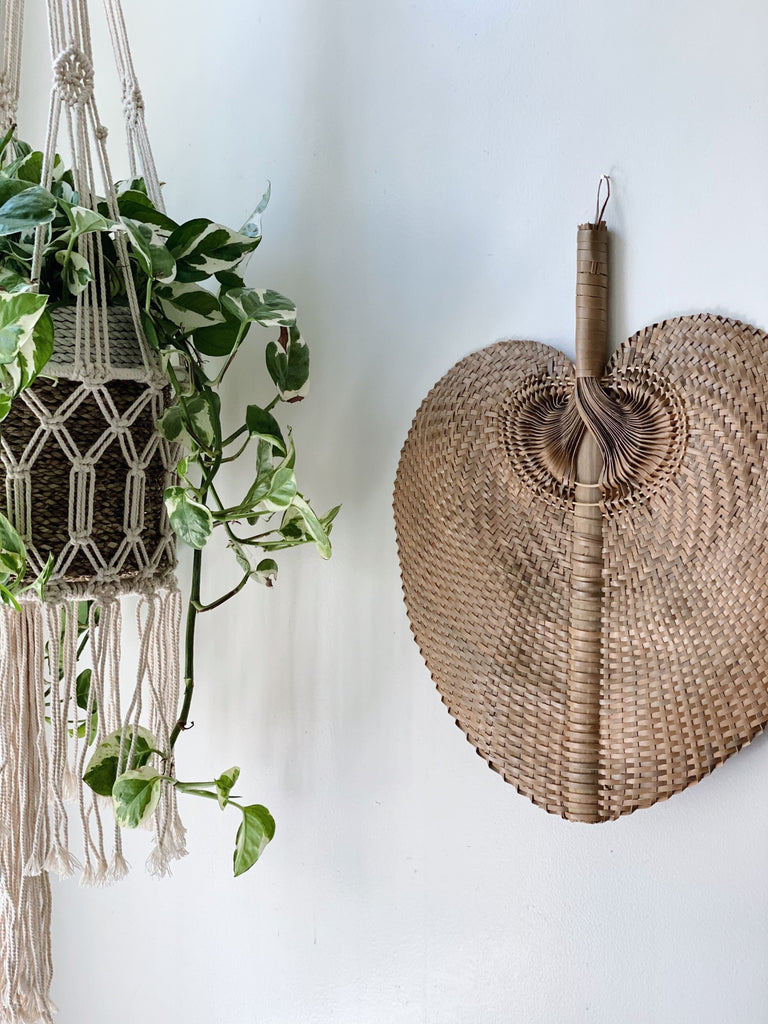 large brown bohemian handwoven palm leaf fan hanging on white wall next to macrame plant hanger with pothos
