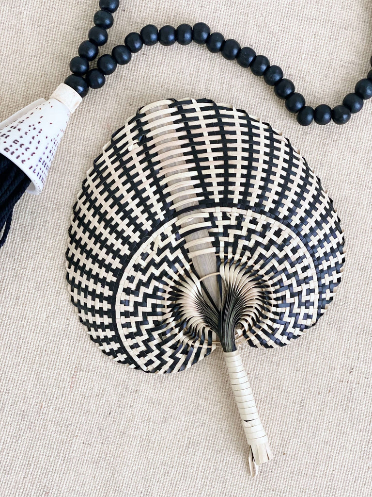 small natural black handwoven traditional design palm leaf fan next to black bohemian wooden bead tassel with beautiful shell