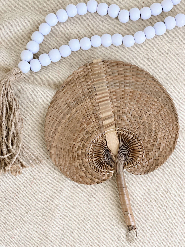 small brown handwoven palm leaf fan next to white and brown bohemian wooden bead tassel