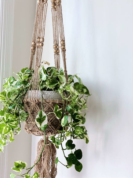 brown beaded jute plant hanger holding a pothos plant in a woven pot against a white wall