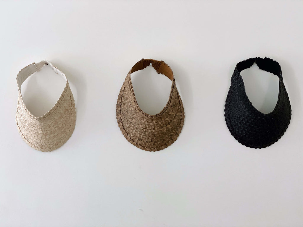 trio of bohemian handwoven white, brown, and black palm leaf sun visors hanging on white wall