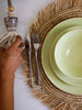 woman setting up table with handwoven straw placemat with sage plates and bohemian shell tassel