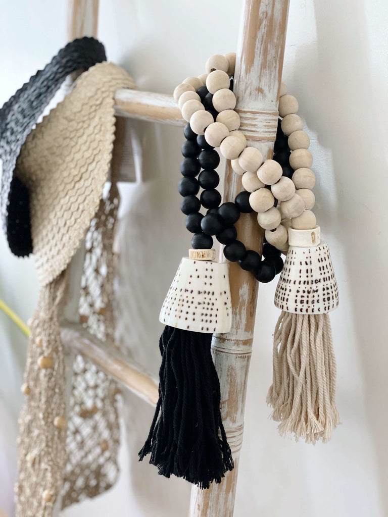 set of two bohemian natural wooden bead shell tassel next to palm leaf visors and jute market bag on bamboo ladder